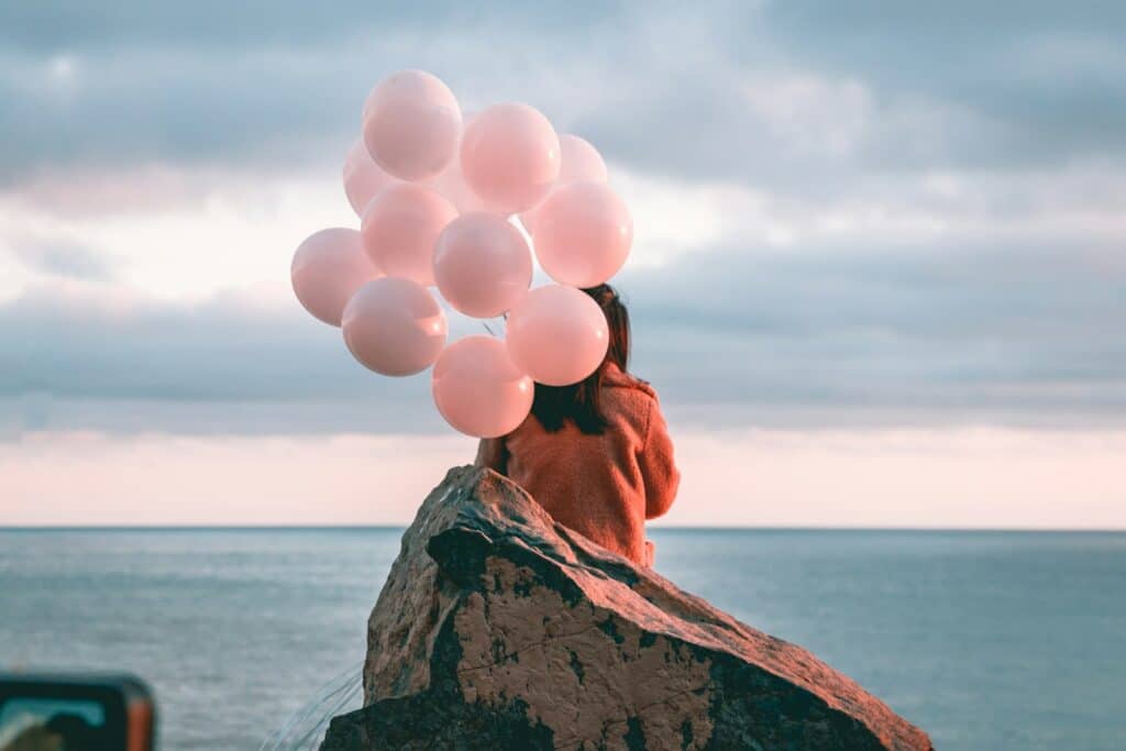 Woman sits with pink balloons overlooking the ocean.